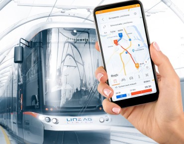 Ein Smartphone mit der Linzmobil App wird in die Kamera gehalten. Im Hintergrund ist eine Straßenbahn zu sehen.