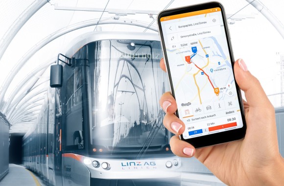 Ein Smartphone mit der Linzmobil App wird in die Kamera gehalten. Im Hintergrund ist eine Straßenbahn zu sehen.