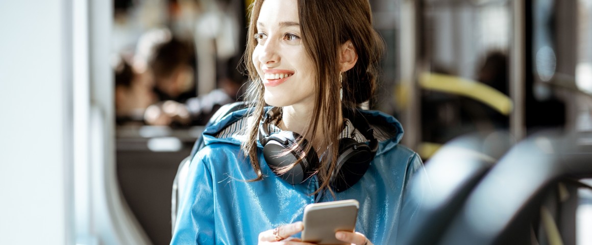 Eine Frau mit einem Handy in der Hand blickt lächelnd aus dem Fenster der Straßenbahn.