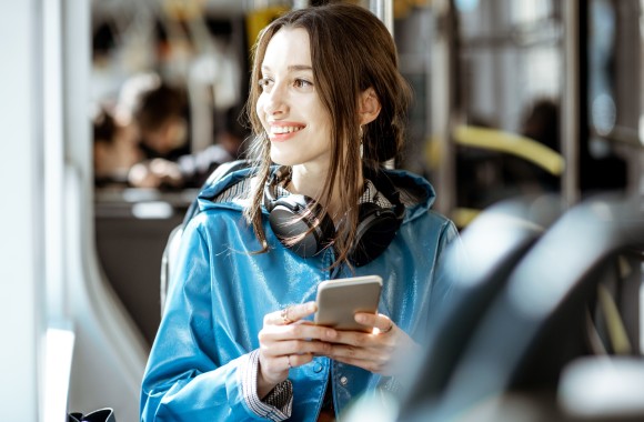 Eine Frau mit einem Handy in der Hand blickt lächelnd aus dem Fenster der Straßenbahn.