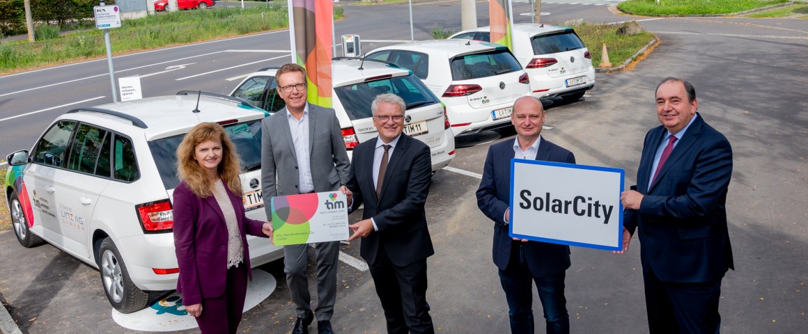 Herr Haider, Waldhör, Lugner, Hein und Frau Rinner stehen mit einem Ortschild der SolarCity vor den Tim Autos.