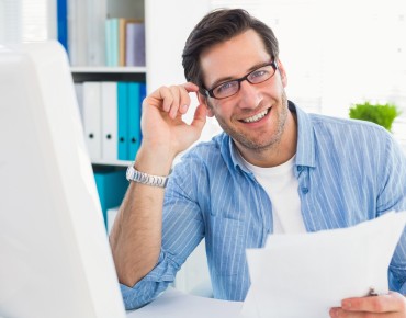 Mann mit Brille sitzt vor PC mit Unterlagen in der Hand und lächelt in die Kamera.