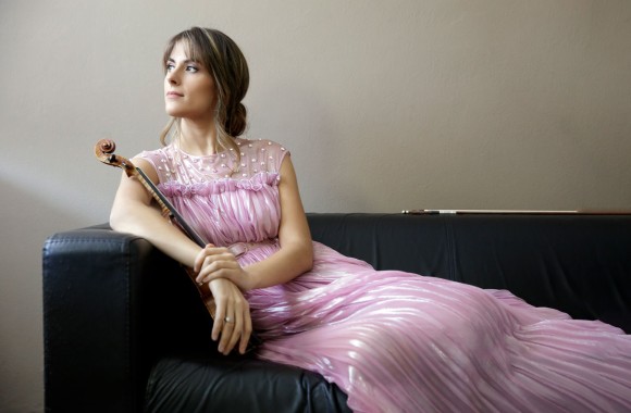 Eine Frau in einen rosafarbenen Kleid lehnt mit einer Geige im Arm auf einer Couch.