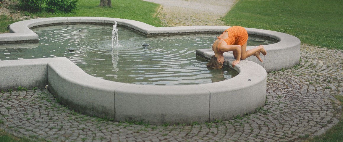 Eine Frau in einem orangefarbenen Sommerkleid kniet kopfüber auf einem Brunnen.