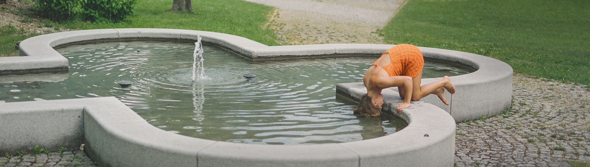 Eine Frau in einem orangefarbenen Sommerkleid kniet kopfüber auf einem Brunnen.