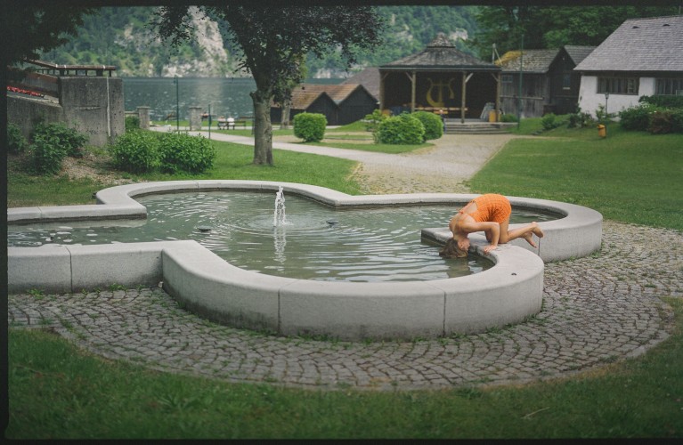 Eine Frau in einem orangefarbenen Sommerkleid kniet kopfüber auf einem Brunnen.