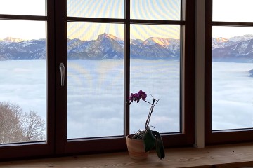 Blick durch ein Fenster auf eine Gebirgskette mit Kaltluftsee davor.