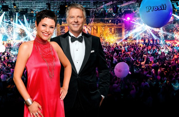 Frau und Mann stehen lächelnd vor der Kamera, im Hintergrund wird eine Silvestershow gefeiert.