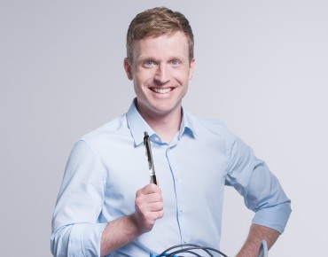 Mann steht lächelnd vor einem Kabelsalat mit einem Stift in der Hand.
