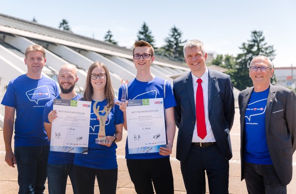 Mehrere Menschen befinden sich vor dem Designcenter in Linz, zwei davon halten einen Pokal und eine Auszeichnung in den Händen.