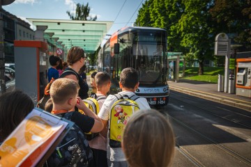 Eine Gruppe von Volksschülern bei der Straßenbahnhaltestelle Wifi/Linz AG beobachtet die Straßenbahn beim Einfahren. Im Vordergrund ist der Aufgabenbogen der Lili Rallye zu sehen.