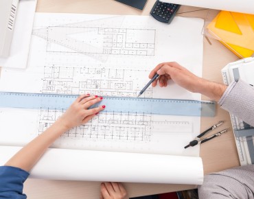 Zwei Personen bearbeiten einen Bauplan mit Lineal und Bleistift.