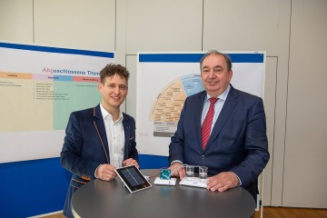 Wasserstoff-Experte in der LINZ AG, Dominik Matheisl, BSc, mit LINZ AG-Generaldirektor DI Erich Haider, davor eine Miniatur einer Wasserstoff-Anlage zur Veranschaulichung, wie klimaneutraler Wasserstoff durch Elektrolyse gewonnen werden kann.