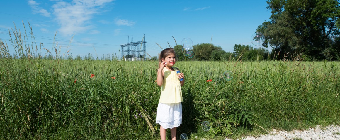 Ein kleines Mädchen spielt mit Seifenblasen am Feldrand, im Hintergrund eine Stromleitung.