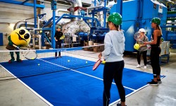 Wättchen spielt Tennis mit zwei Frauen und einem Mann in der Turbinenhalle des Fernheizkraftwerkes.