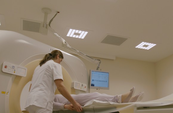 Eine Frau schiebt eine andere Person in das MR Gerät in einer Arztpraxis. Der Raum ist hell mit LED Beleuchtung ausgeleuchtet