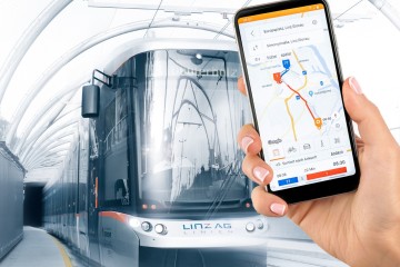 Bild von einem Smartphone an welchem die Linz-Mobil-App geöffnet ist. Im Hintergrund ist eine Straßenbahn zu sehen.