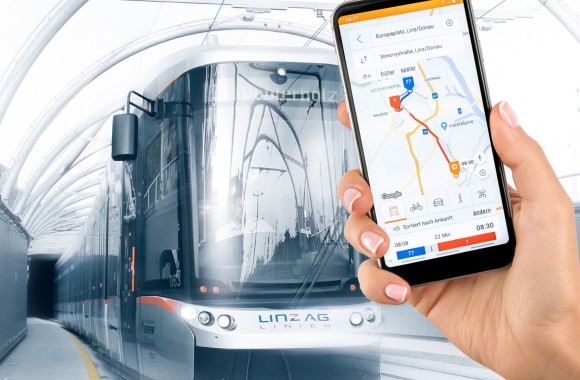 Bild von einem Smartphone an welchem die Linz-Mobil-App geöffnet ist. Im Hintergrund ist eine Straßenbahn zu sehen.
