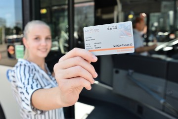 Frau steht lächelnd vor Straßenbahn und hält das Mega-Ticket in die Kamera