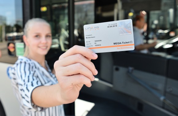 Frau steht lächelnd vor Straßenbahn und hält das Mega-Ticket in die Kamera