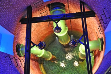 Bild des Schaubrunnens im Wasserwerk Scharlinz. Man sieht einen runden Brunnenschacht, blau beleuchtet und grüne Wasserrohre die bis zum Grund des Brunnens reichen. Am unteren Ende des Brunnenschachts ist Wasser zu erkennen.