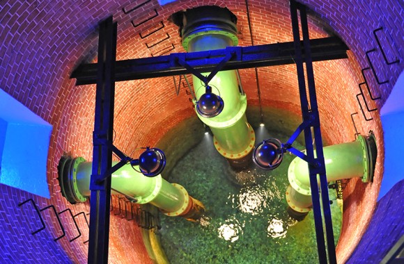 Bild des Schaubrunnens im Wasserwerk Scharlinz. Man sieht einen runden Brunnenschacht, blau beleuchtet und grüne Wasserrohre die bis zum Grund des Brunnens reichen. Am unteren Ende des Brunnenschachts ist Wasser zu erkennen.