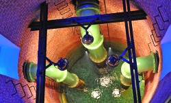 Bild des Schaubrunnens im Wasserwerk Scharlinz. Man sieht einen runden Brunnenschacht, blau beleuchtet und grüne Wasserrohre die bis zum Grund des Brunnens reichen. Am unteren Ende des Brunnenschachts ist Wasser zu erkennen.