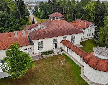 Das Wasserwerk Scharlinz, gelegen inmitten des Linzer Wasserwalds, von oben. Weißes Gebäude mit rotem Dach inmitten von grünen Wiesen und umgeben von Bäumen.