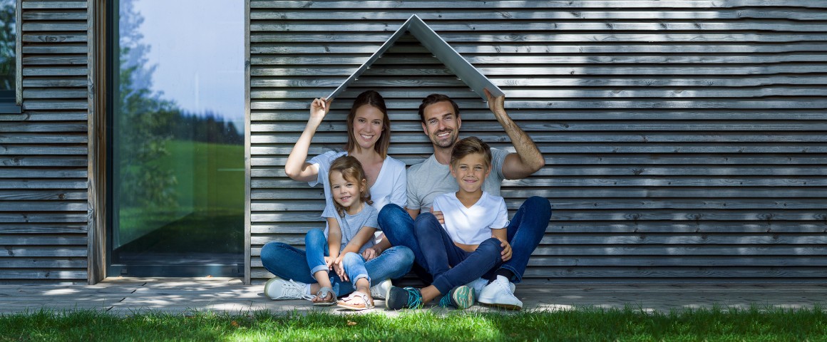 Mutter, Vater und ihre zwei Kinder sitzen unter einem aus weißem Karton gefaltenen Dach am Boden vor einer weißen Mauer.