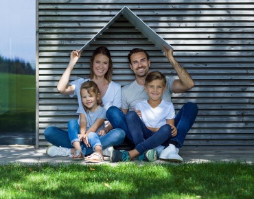 Mutter, Vater und ihre zwei Kinder sitzen unter einem aus weißem Karton gefaltenen Dach am Boden vor einer weißen Mauer.