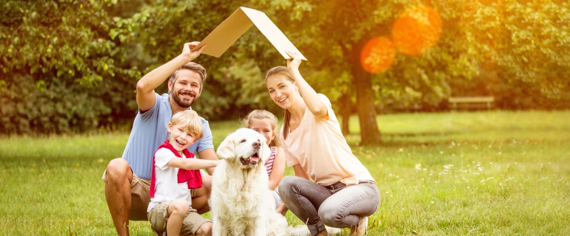 Eine Familie sitzt auf einer Wiese und hält sich dabei ein symbolisches Dach über den Kopf.