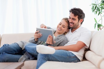 Ein junges Pärchen sitzt aneinander geschmiegt auf der Couch, beide lächeln und schauen in ein Tablet