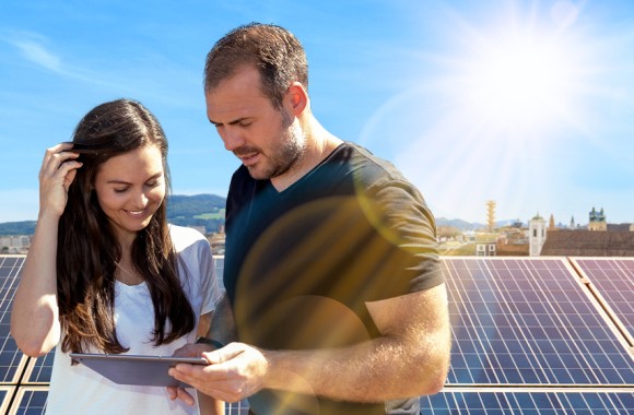 Eine Frau und ein Mann sitzen vor einer Photovoltaikanlage und schauen gemeinsam in ein Tablet.