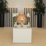 Hölzerner Sarg steht in einem hellem Raum auf einem weißen Podest. Im Hintergrund sind lange schmale schwarze Kerzen, links und rechts davon eine hohe Plfanze.