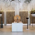 Hölzerner Sarg steht in einem hellem Raum auf einem weißen Podest. Links und rechts davon sind Dekorationselemente mit natürlichem Grün. Im Hintergrund stehen an der Wand lange schmale schwarze Kerzen. An der Decke sind Äste angebracht, welche herunterhängen.