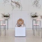 Sarg steht in einem hellen Raum auf weißem Podest. An der Decke hängen Äste herunter. Links und rechts neben dem Sarg sind Dekoschüssel gefüllt mit Ästen.