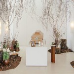 Hölzerner Sarg steht auf weißem Podest in einem Raum mit weißen Wänden. Links und rechts sind Waldböden aufgestreut mit abgeschnittenen Baumstämmen und hohen Birken. Auf jedem Baumstamm stehen Kerzen. Vor dem Sarg auf dem Podest ist ein Foto des*der Verstorbenen.