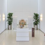 Hölzerner Sarg steht in hellem, weiß gestrichenen Raum auf weißem Podest. Foto des*der Verstorbenen und weiße Orchideen vor dem Sarg. Hinten in den beiden Ecken stehen grüne Pflanzen und weiße lange brennende Kerzen. Auf der rechten Seite an der Wand stehen noch zwei rote Stühle.