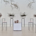 In der Mitte steht die Urne, daneben ein Foto des*der Verstorbenen. Heller weiß gestrichener Raum in dem vier Dekoschüsseln mit Ästen dekoriert. Von der Decke hängen Äste herunter.