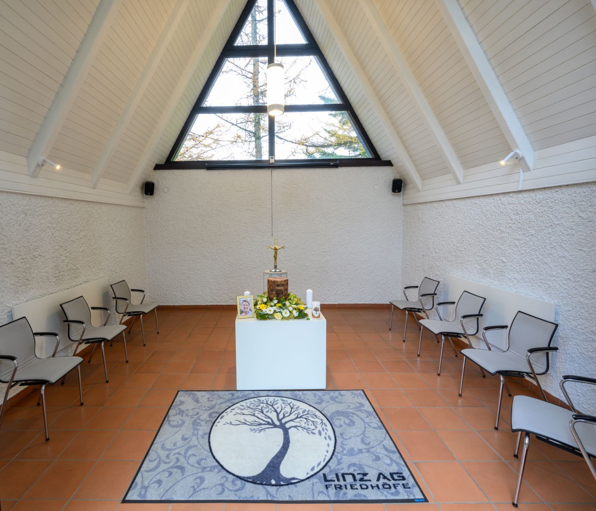 Schwarze Urne steht in einem hellem Raum. Links und rechts davon sind Dekorationselemente mit natürlichem Grün. Im Hintergrund stehen an der Wand lange schmale schwarze Kerzen. An der Decke sind Äste angebracht, welche herunterhängen. Im Vordergrund drei goldene, dekorierte Podeste.