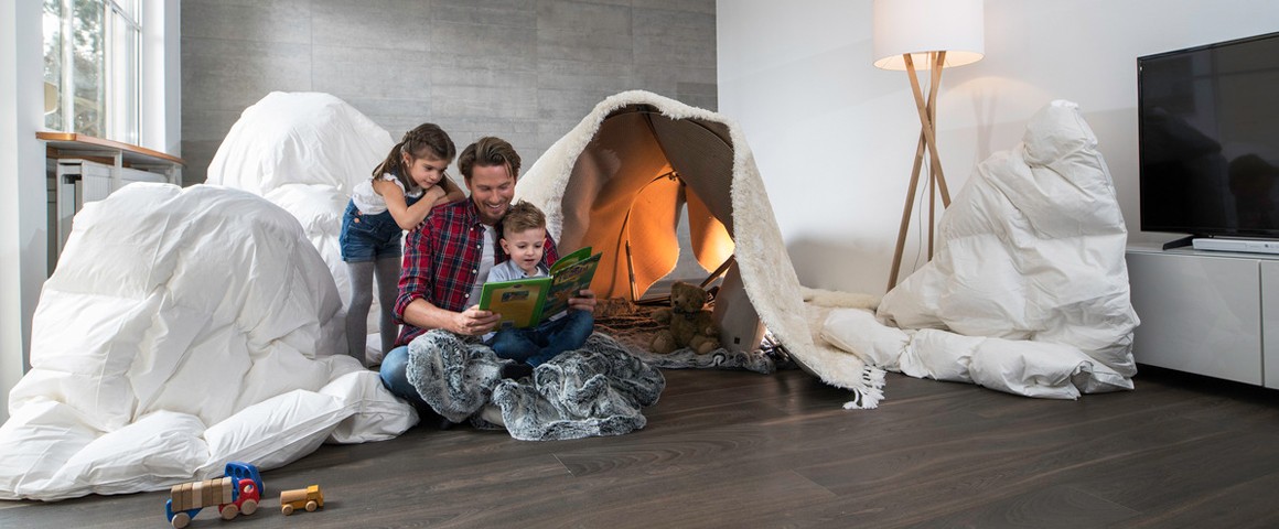 Ein Vater sitzt mit seinen beiden Kindern im Wohnzimmer am Boden und blättert einen Bildband durch. Im Hintergrund ein selbstgebautes Zelt aus Bettlaken und Decken.