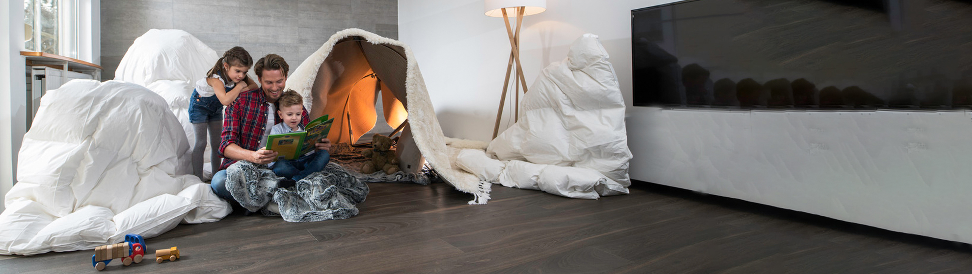 Ein Vater sitzt mit seinen beiden Kindern im Wohnzimmer am Boden und blättert einen Bildband durch. Im Hintergrund ein selbstgebautes Zelt aus Bettlaken und Decken.