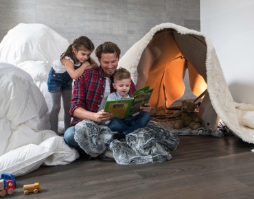 Ein Vater sitzt mit seinen beiden Kindern im Wohnzimmer am Boden und blättert einen Bildband durch. Im Hintergrund ein selbstgebautes Zelt aus Bettlaken und Decken.