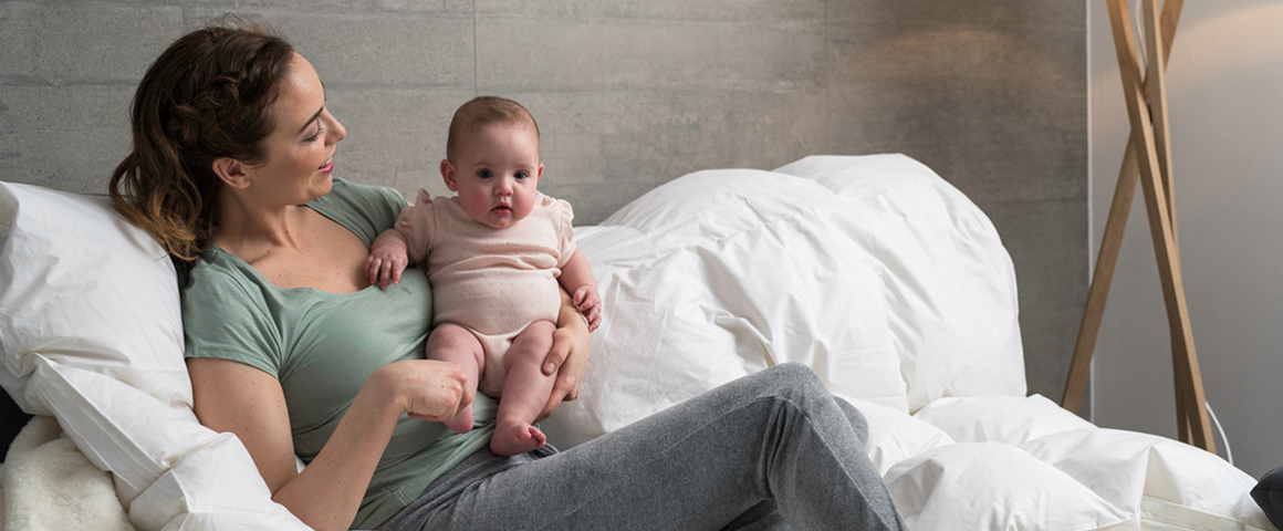 Eine junge Mutter sitzt mit ihrem Baby im Arm am Bett und lächelt es an.