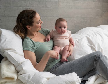 Eine junge Mutter sitzt mit ihrem Baby im Arm am Bett und lächelt es an.