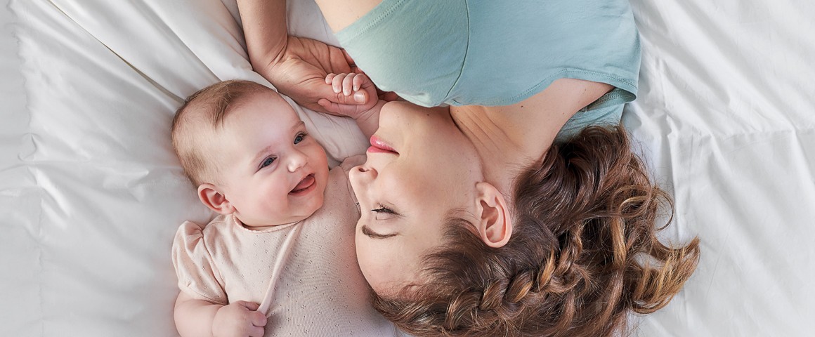 Eine Frau liegt mit ihrem Baby im Bett, sie lächeln sich an