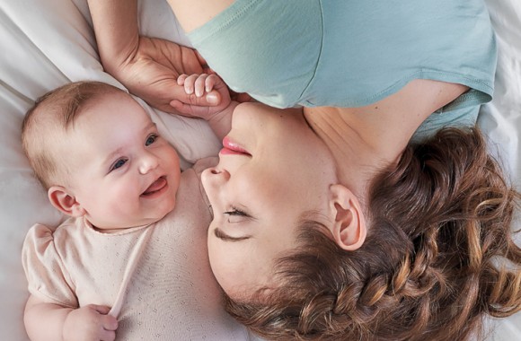 Eine Frau liegt mit ihrem Baby im Bett, sie lächeln sich an