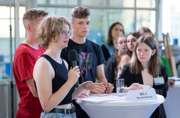 Jugendliche stehen um einen runden Tisch. Ein Mädchen hält ein Mikrofon in der Hand und stellt eine Frage.
